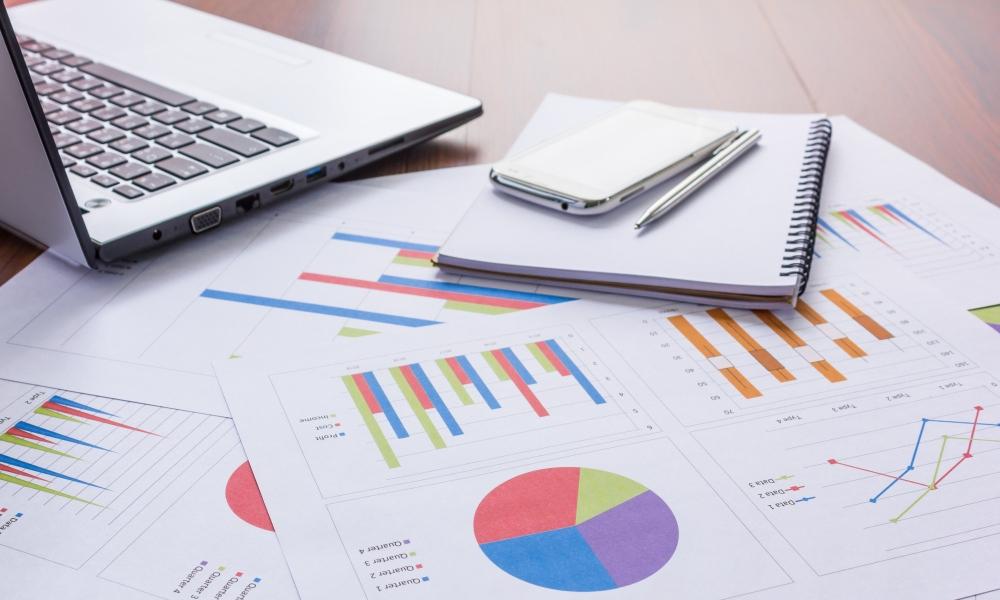 Laptop and notebook placed on printed reports with bar charts, pie charts, and data visualizations.